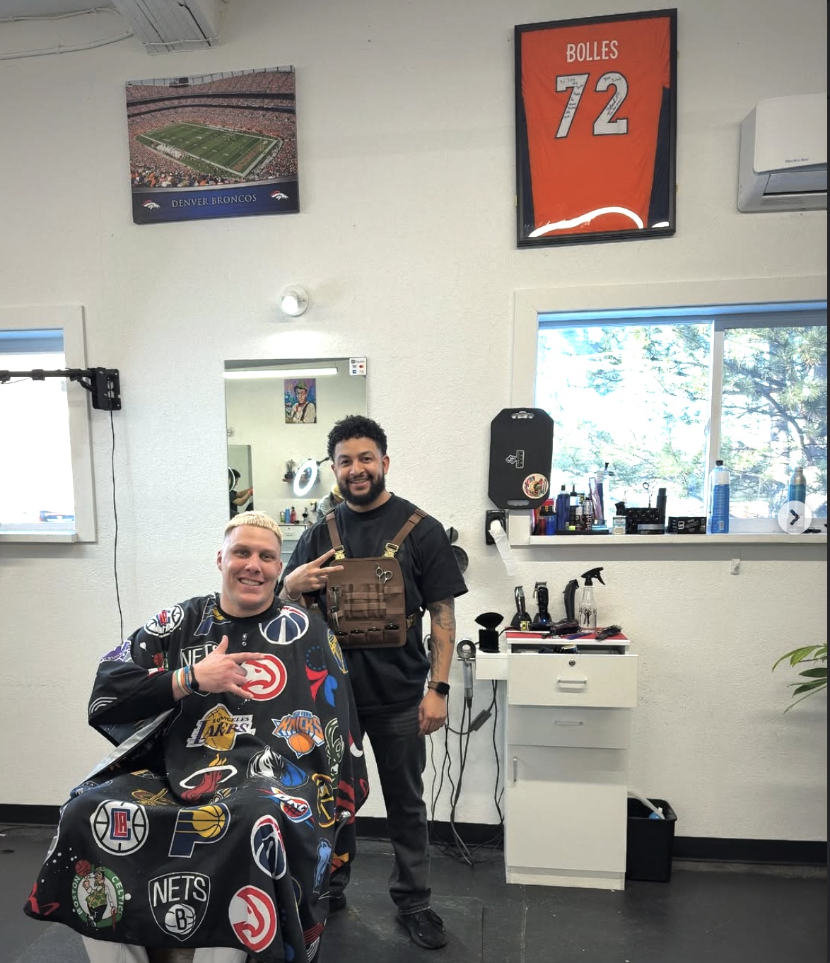 Owner Jose Barraza in Cultura Barbershop, with a framed Denver Broncos jersey and stadium poster on the wall behind him